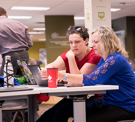 Two women sitting at a desk in a room full of people
