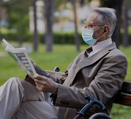 Older adult man sitting on a park bench reading the newspaper with trees and grass in the background